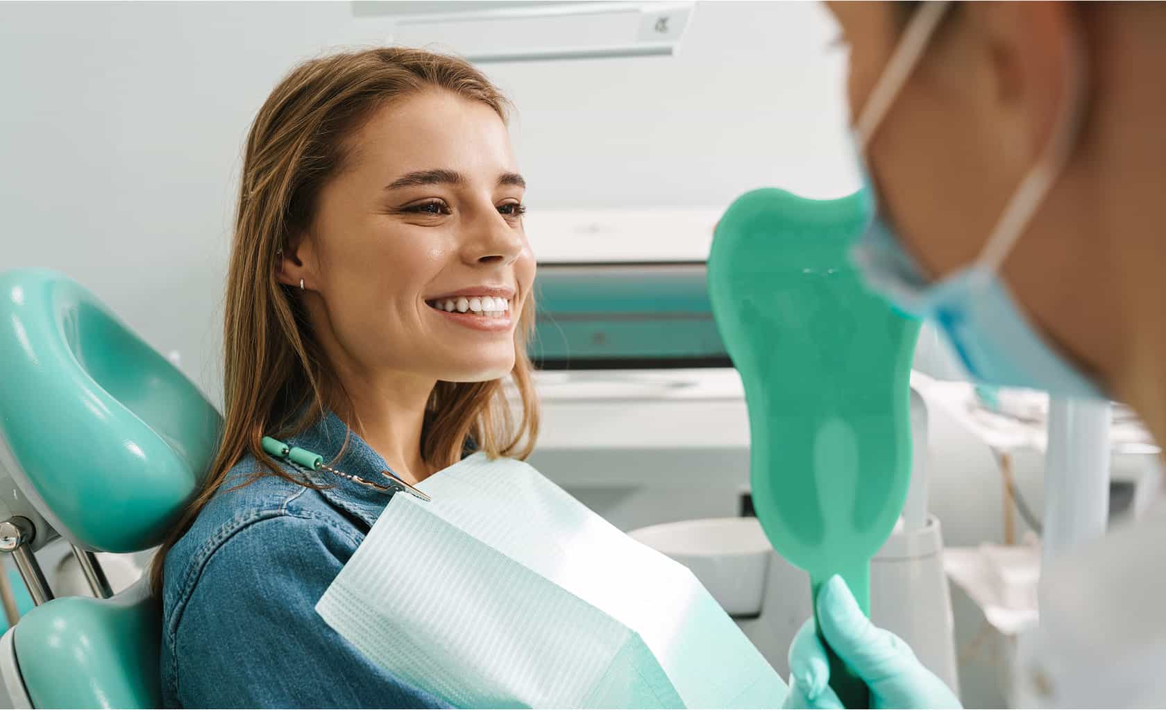 Smiling young woman in dental chair looking at her teeth with dentist holding a mirror during check-up